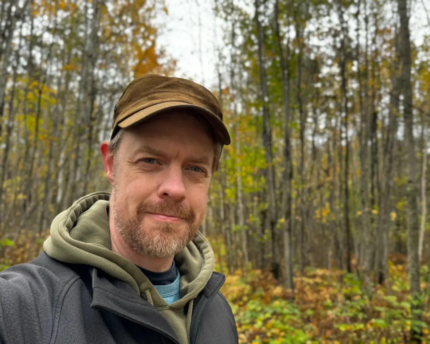 Selfie of the author in the woods, wearing a green cap and a hoodie under a black jacket